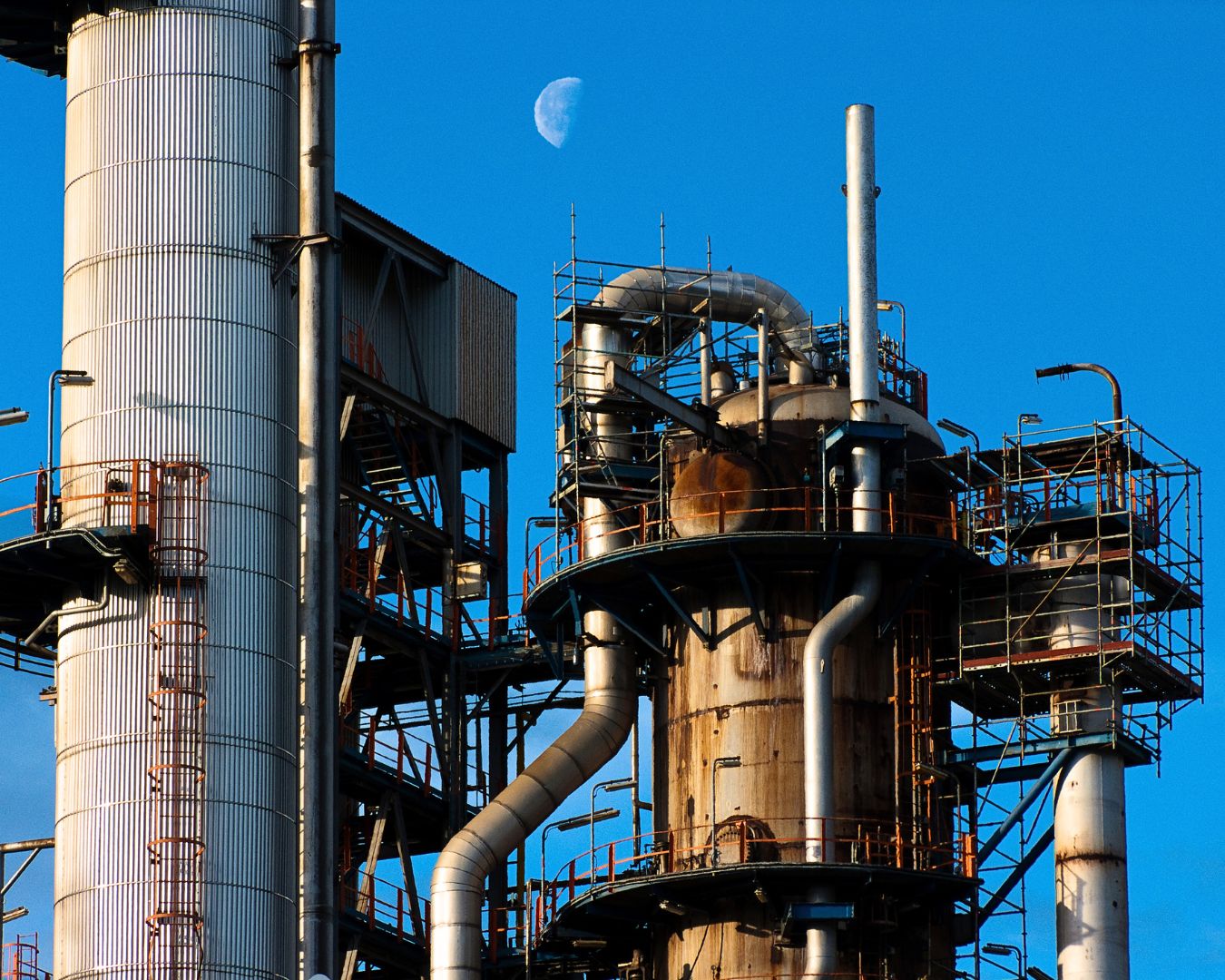 Planta industrial del País Vasco en funcionamiento, con estructuras metálicas y tuberías, destacando la producción bajo un cielo azul con la luna visible.